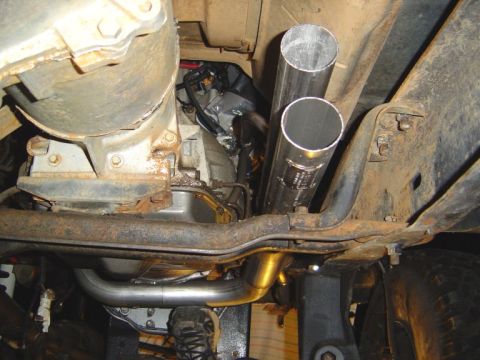 1986 Ford Bronco - Underside view of a vehicle showing two shiny stainless-steel exhaust pipes routed vertically near a black frame rail, with a rusty differe…