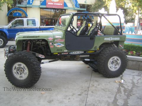 1990 Ford Bronco - green Jeep-bodied rock crawler with full roll cage, BFGoodrich tires and front winch parked on a plaza walkway.