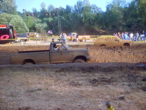 1976 Ford F-100 - Multiple vehicles are stuck in a mud pit during an event, with spectators in the background.