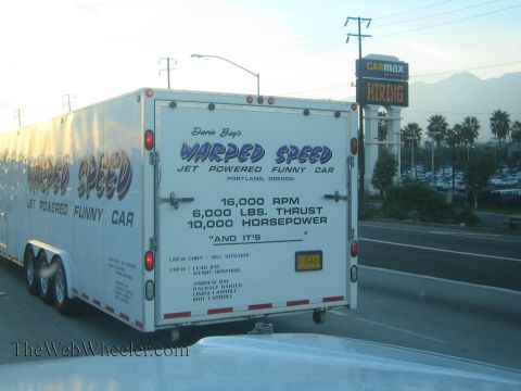 1990 Ford Bronco - Rear of a white enclosed trailer reading Warped Speed Jet Powered Funny Car, photographed on a highway near a CarMax sign.