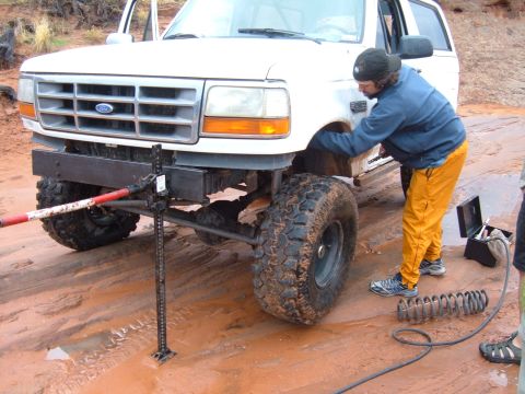 1993 Ford Bronco - A person is using a Hi-Lift jack to lift a modified Ford Bronco in a muddy, off-road environment.