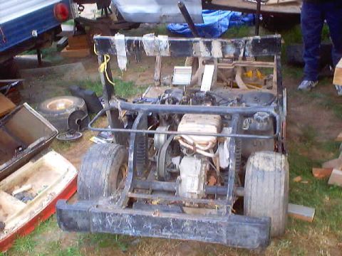 1976 Ford F-100 - A stripped, dust-covered go-kart frame sits on a grassy yard, viewed from the rear with its tube roll cage, exposed single-cylinder engine,…