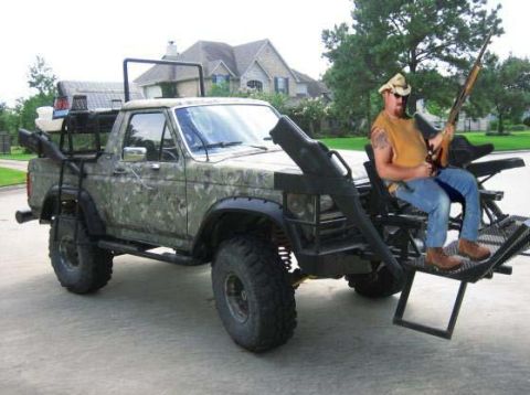 1989 Ford Bronco - A man sits in a hunting stand attached to the rear of a camouflaged pickup truck, holding a rifle.