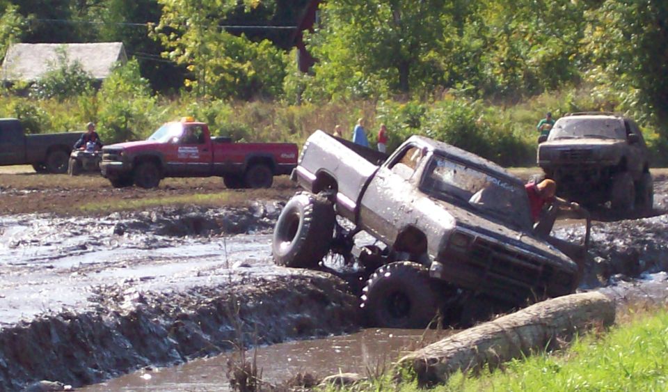 Muddy pickup truck with large tires stuck in a bog, another truck and ATV visible