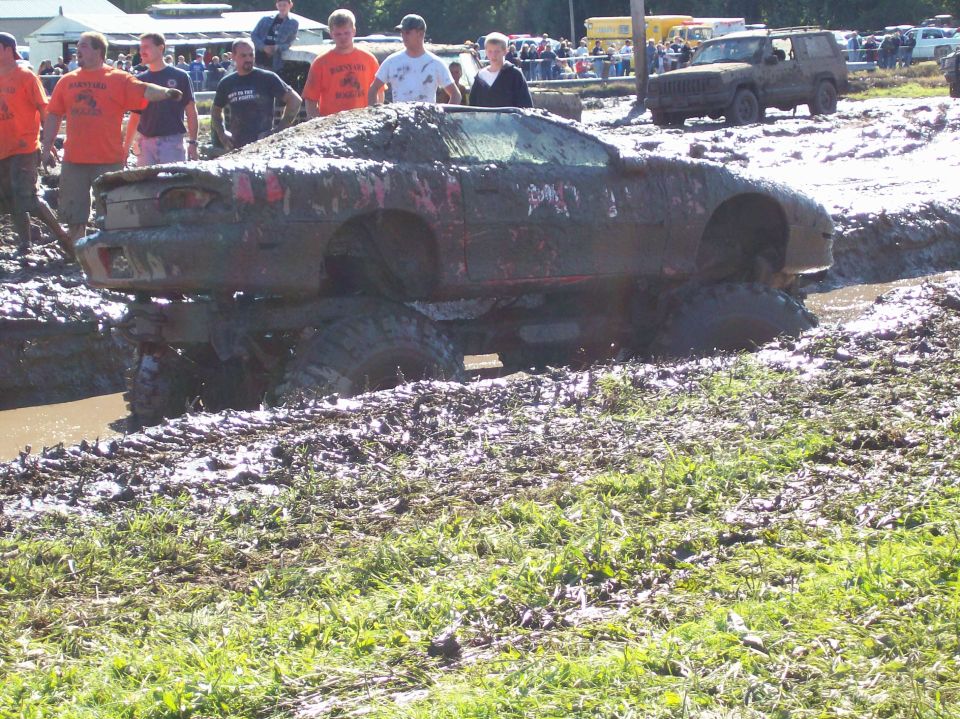 Mud-covered red car stuck in a mud pit with large tires visible