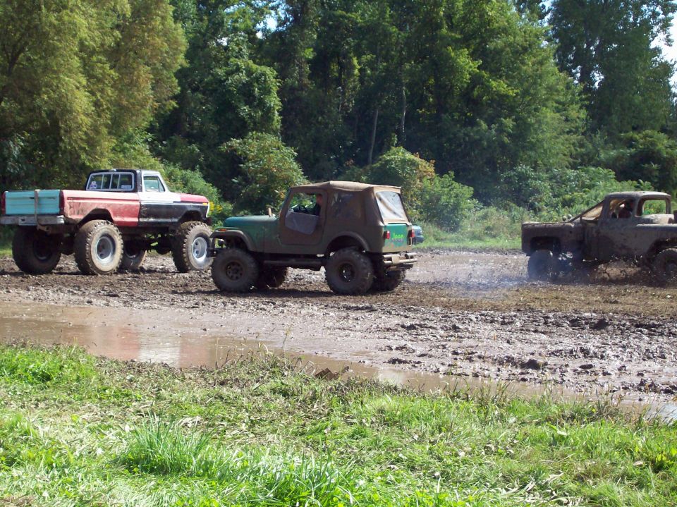 Three modified off-road trucks and SUVs are driving through a muddy track with trees in the background