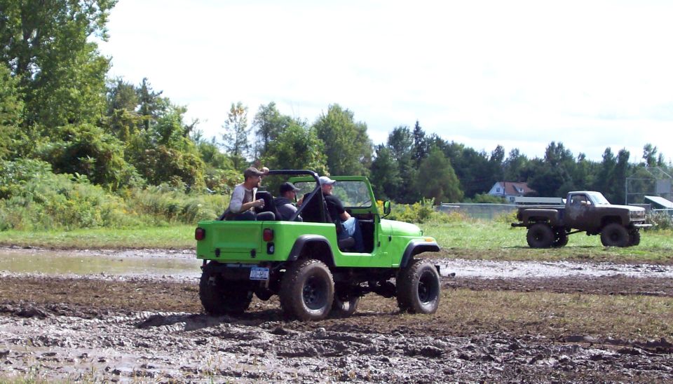 Green Jeep Wrangler with three people driving through mud, a brown pickup truck in the background