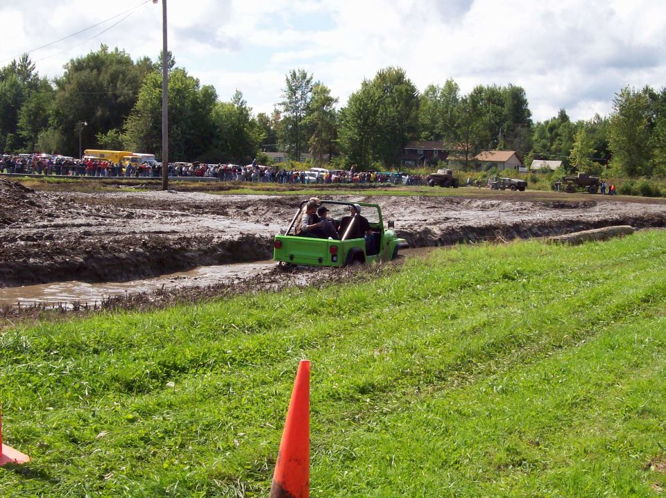 Green off-road vehicle navigating a mud pit with spectators in the background