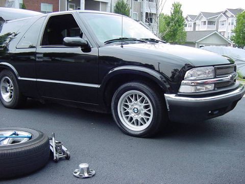 1984 BMW 3 Series - A black Chevrolet Blazer is parked on asphalt with a flat tire, a jack, and a spare tire nearby.