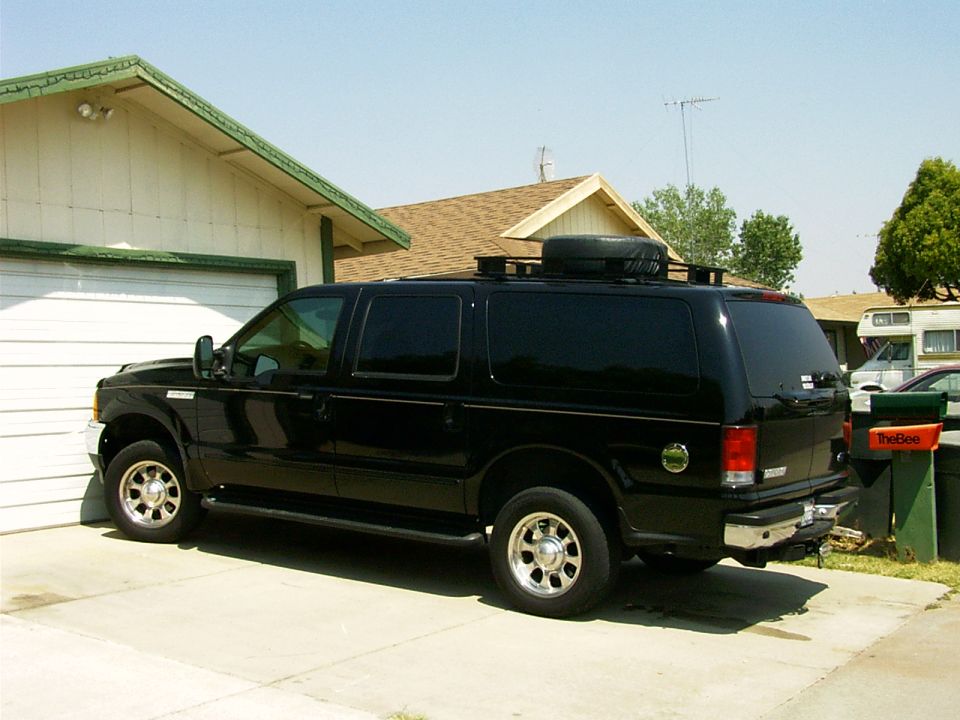 Black Ford Excursion SUV parked in driveway, side/rear view with roof rack and spare tire.