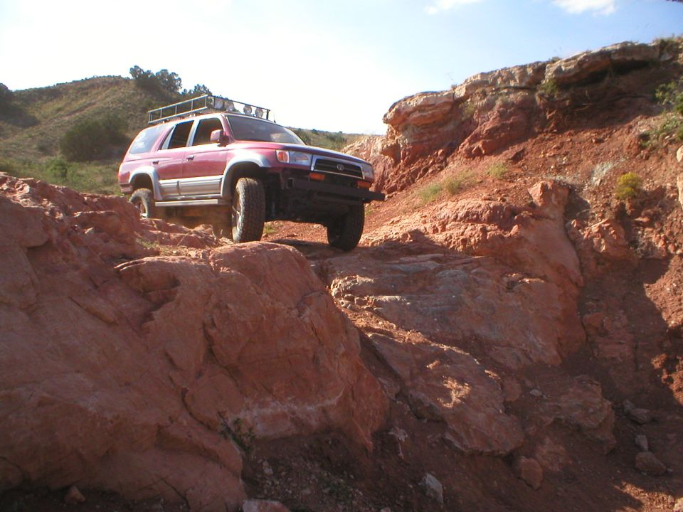 Red 1996 Toyota 4Runner with roof rack driving on a rocky, off-road trail