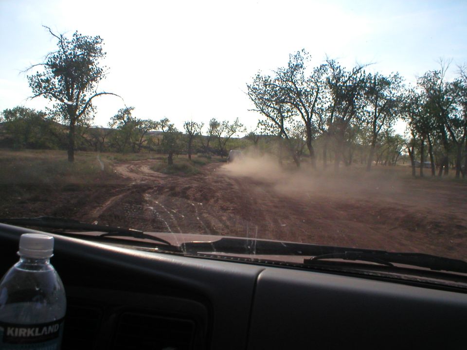 View from inside a vehicle driving on a dirt road, kicking up dust. Trees and dry brush line the path