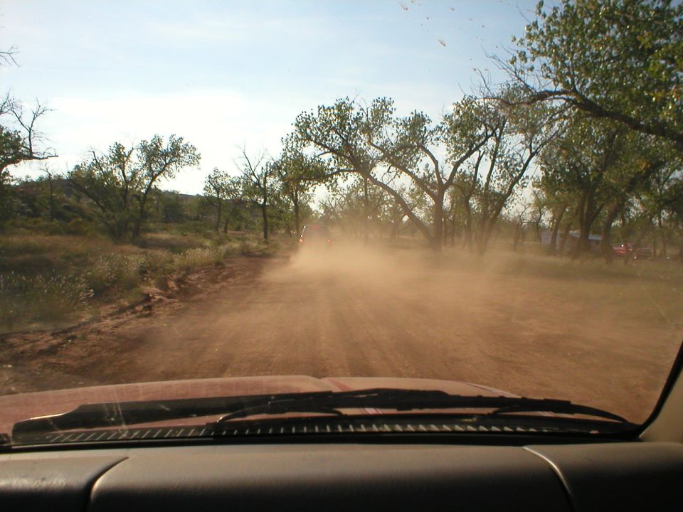 Driving down a dirt road kicking up dust, trees on the side, other vehicles visible ahead