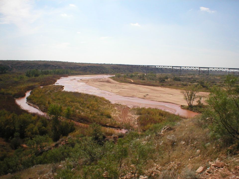 Wide view of a muddy river flowing through a dry landscape with a long bridge in the distance