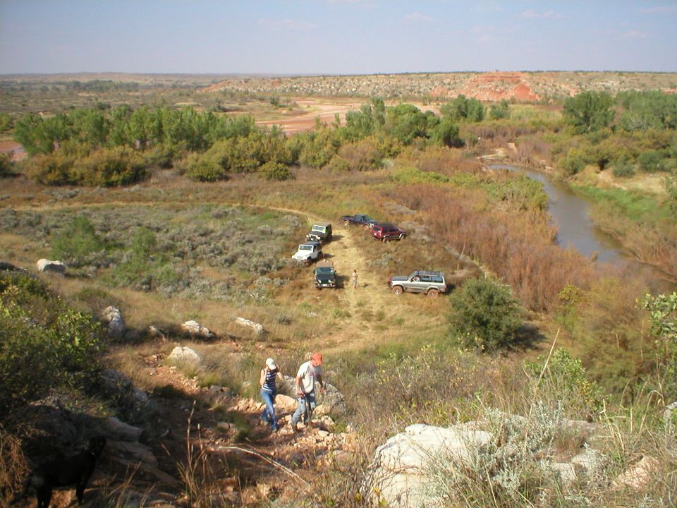 Several SUVs parked on a dirt path next to a river, with two people walking down a rocky hill