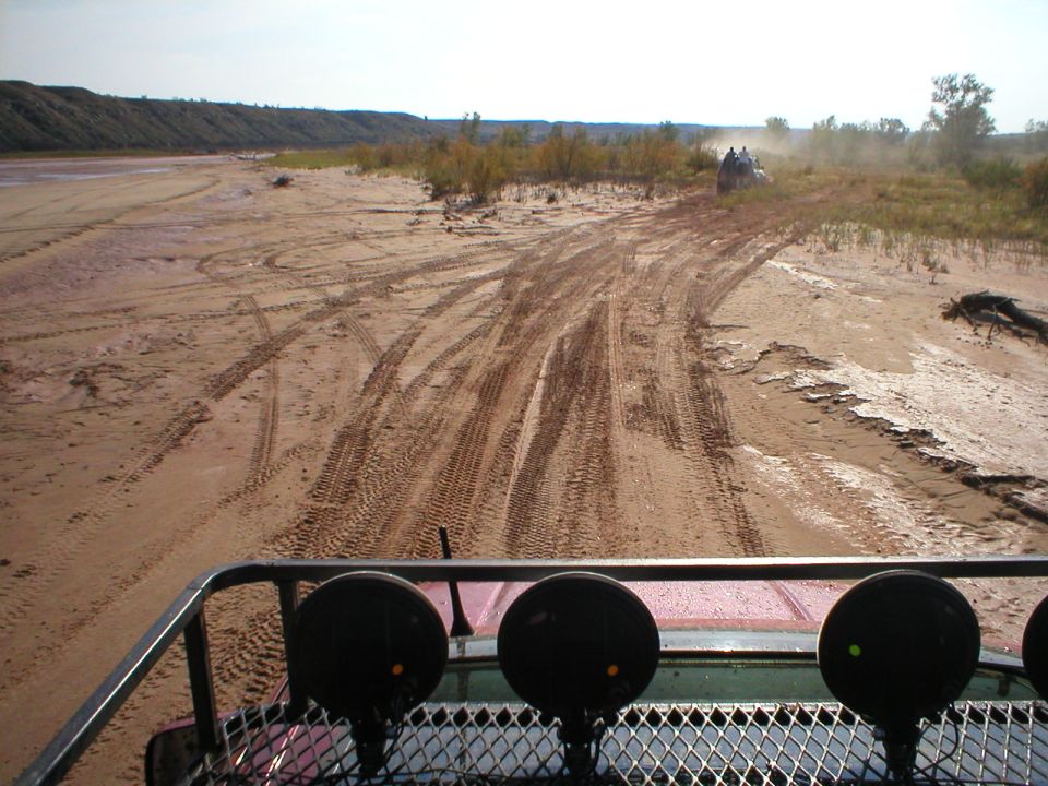 View from a vehicle looking at tire tracks in sand, with other vehicles in the distance