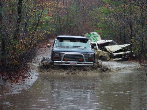 1978 Ford Bronco - A blue Ford Bronco navigates a flooded path, with a damaged vehicle partially visible behind it.