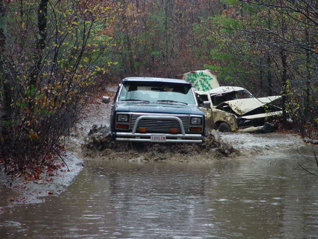 Blue 1980s Ford Bronco driving through muddy water, front view