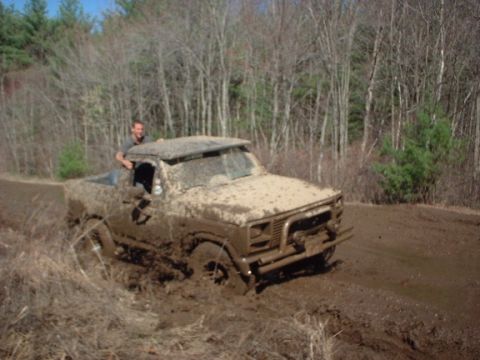 1978 Ford Bronco - Truck stuck deep in mud on a wooded trail, driver visible.