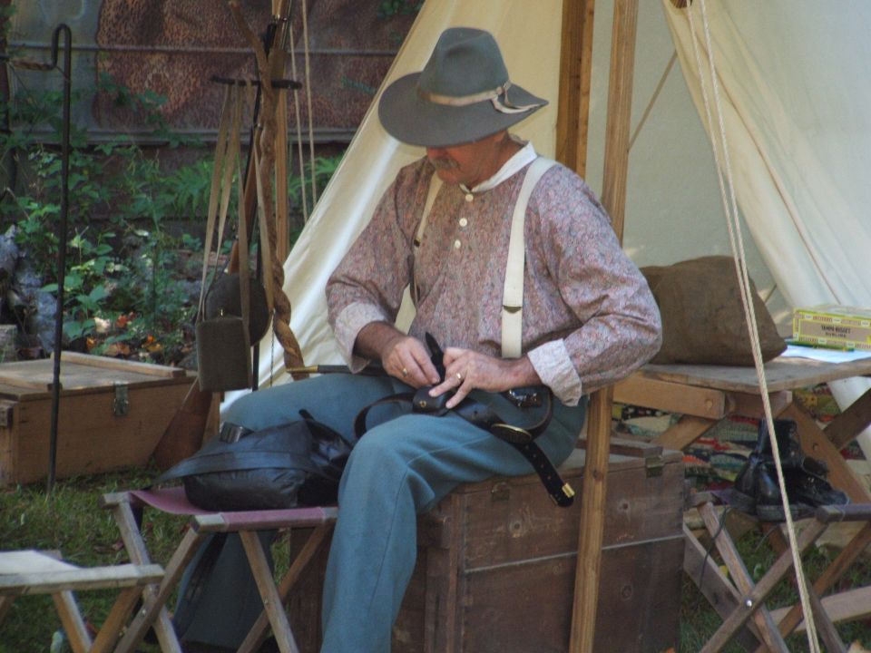 Man in historical reenactment attire sits near a tent, adjusting a belt