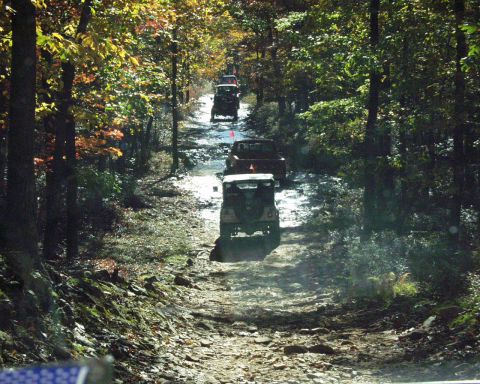 1989 Ford Bronco - Multiple off-road vehicles navigate a challenging dirt path surrounded by trees in autumn foliage.