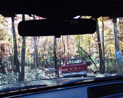 1989 Ford Bronco - A red Ford pickup truck is seen from inside another vehicle, driving through a wooded area.
