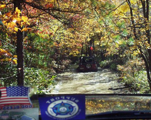 1989 Ford Bronco - Jeep Wrangler navigates a dirt road surrounded by colorful autumn trees.