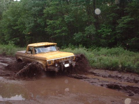 1978 Ford Bronco - A yellow pickup truck navigates a muddy track, kicking up a large spray of mud.