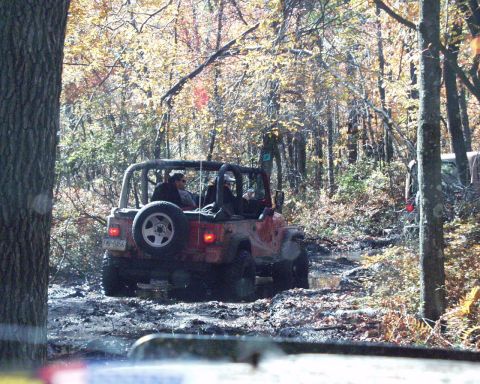 1989 Ford Bronco - Two off-road vehicles navigate a challenging muddy path in a wooded area during autumn.