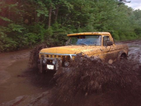 1978 Ford Bronco - A yellow pickup truck navigates a muddy terrain, kicking up a large spray.