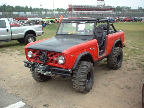 1978 Ford Bronco - A modified red SUV with large off-road tires and a winch is parked on a dirt lot, with a stadium in the background.