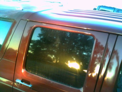 1988 Ford C-350 (Centurion Conversions) - The side profile of a red van shows its window reflecting trees and the setting sun. Another vehicle is parked behind it.