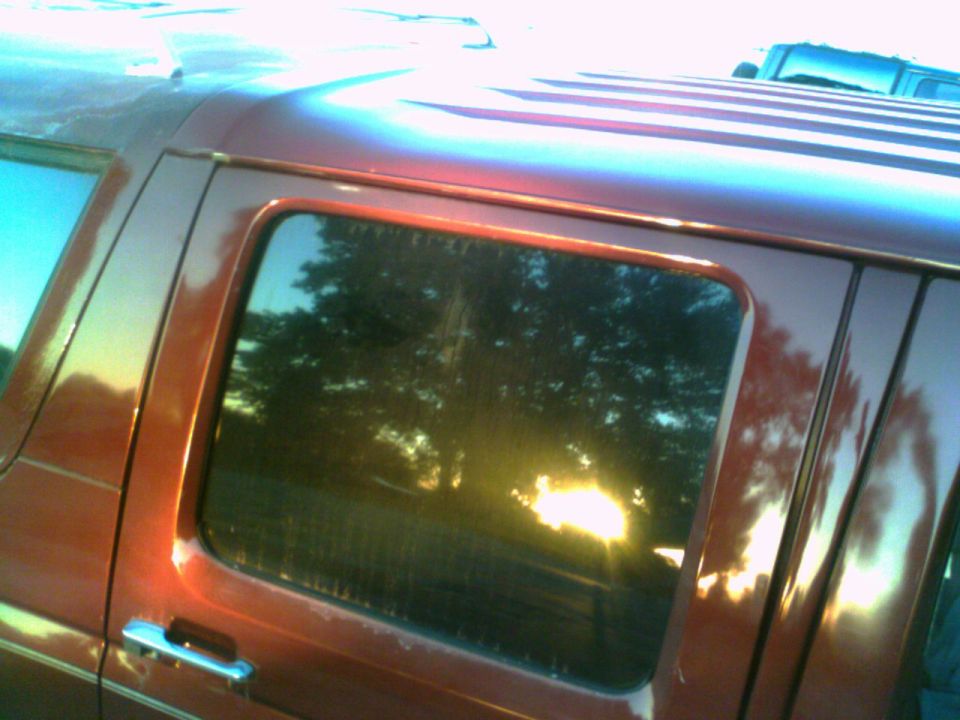 Red van side window reflecting trees and sun. Another vehicle is visible in the background