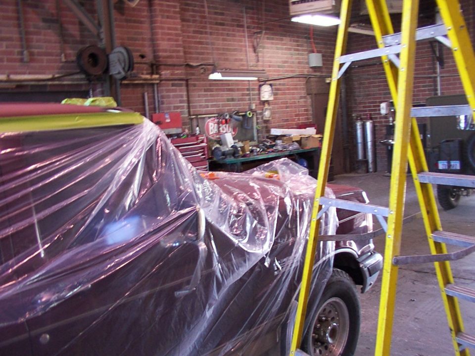 Maroon vehicle covered in plastic sheeting in a garage with a yellow ladder nearby