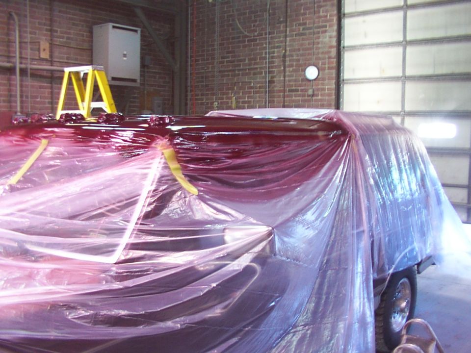 Vehicle covered in plastic sheeting, undergoing painting in a garage. A ladder is visible nearby