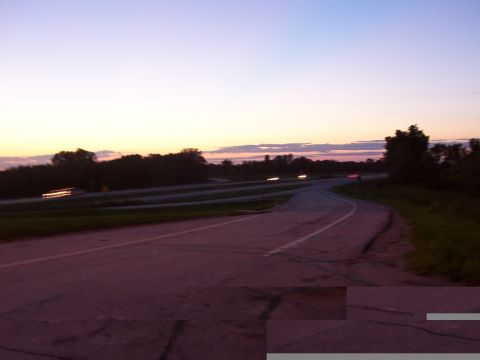 1988 Ford C-350 (Centurion Conversions) - Dusk settles over a highway interchange, with trees lining the horizon and faint car lights in the distance.