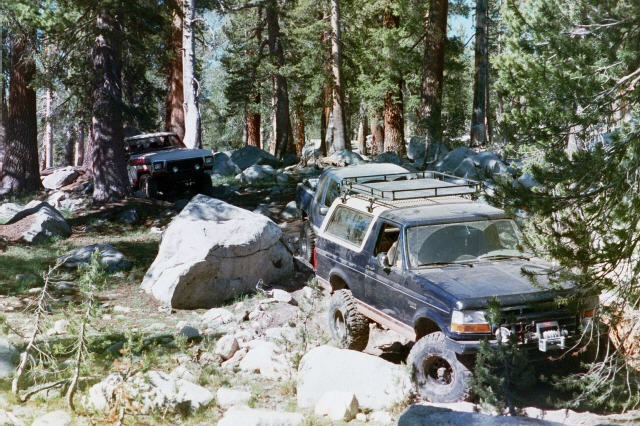 Lifted dark blue Ford Bronco with roof rack and winch crawling over rocks on a forest trail, another SUV behind