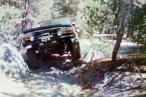 1994 Ford Bronco - A Ford Bronco is shown tackling a challenging off-road path with large rocks and trees surrounding it.