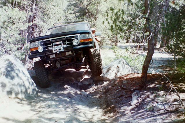 Dark Ford Bronco flexing over rocks on a forested off-road trail with front wheel lifted.
