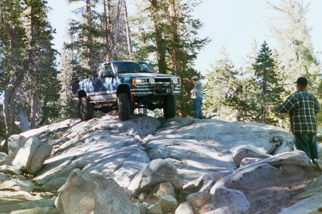 Green lifted 4x4 crawling over granite rocks with two spotters, pine forest behind.