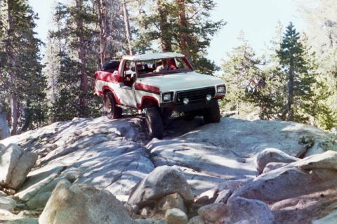 1994 Ford Bronco - Off-road pickup truck tackling a challenging rocky path in a wooded area.