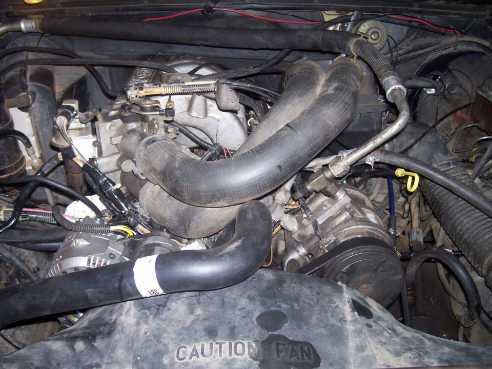 Engine bay with hoses, belts, and alternator visible. Fan shroud has 'CAUTION FAN' text