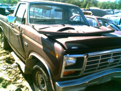 1988 Ford C-350 (Centurion Conversions) - This brown Ford pickup truck is parked in a junkyard with other vehicles visible in the background.