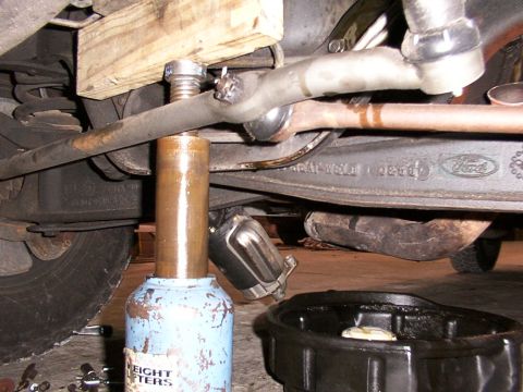 1988 Ford C-350 (Centurion Conversions) - A jack is positioned under a tie rod assembly, with an oil drain pan visible beneath the vehicle's undercarriage.