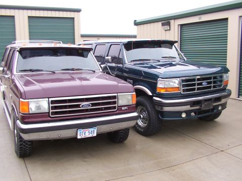1988 Ford C-350 (Centurion Conversions) - Two Ford trucks, a purple model and a dark green model, are parked side-by-side in front of storage unit doors.