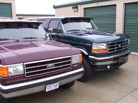 1988 Ford C-350 (Centurion Conversions) - Two Ford trucks are parked side-by-side in front of storage unit doors. The green truck appears to be from the early 1990s.