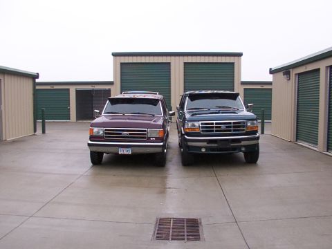 1988 Ford C-350 (Centurion Conversions) - Two Ford trucks are parked in front of storage units. The blue truck's grille suggests a 1992 model year.