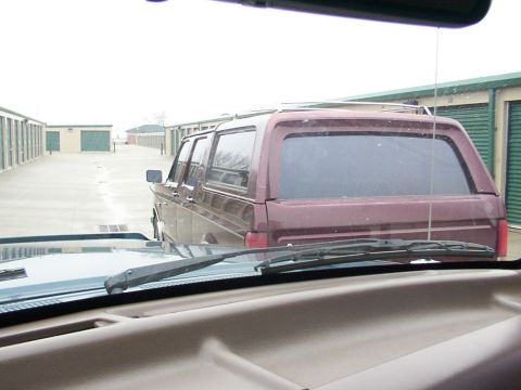 1988 Ford C-350 (Centurion Conversions) - View from inside a vehicle looking out at a maroon SUV with a camper shell parked in a storage unit facility.