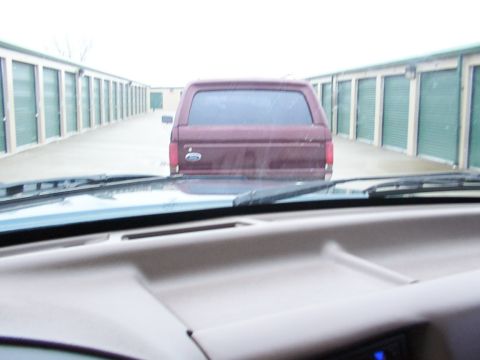 1988 Ford C-350 (Centurion Conversions) - A maroon pickup truck is seen from inside another vehicle, parked in a row of storage units.