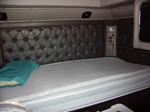 1988 Ford C-350 (Centurion Conversions) - The interior of a truck sleeper cab features a large bed with a teal pillow and a dark, tufted headboard. Control panel visible on the righ…